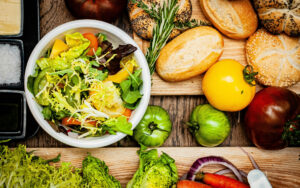 A colorful food spread with a fresh salad in a white bowl filled with leafy greens, tomatoes, and peppers. Surrounding the bowl are assorted bread rolls with seeds, herbs like rosemary, yellow and green tomatoes, red onions, carrots, and lettuce, all arranged on a rustic wooden table.