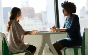Two women, a counsellor and client, sitting across from each other at a small table by a window, engaged in conversation. One woman wears a beige blouse, the other wears a blue long-sleeve top and glasses. Green chairs frame the scene, with a bright cityscape visible through the window behind them.