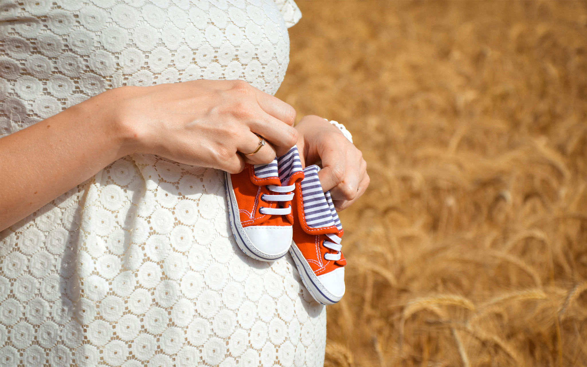 A woman in a white dress holding red baby booties against her stomach.